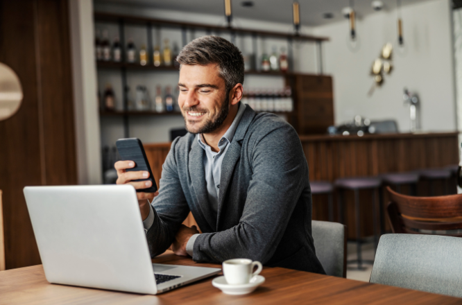 Joven trabajando frente a una laptop sonriendo y con un movil en la mano.