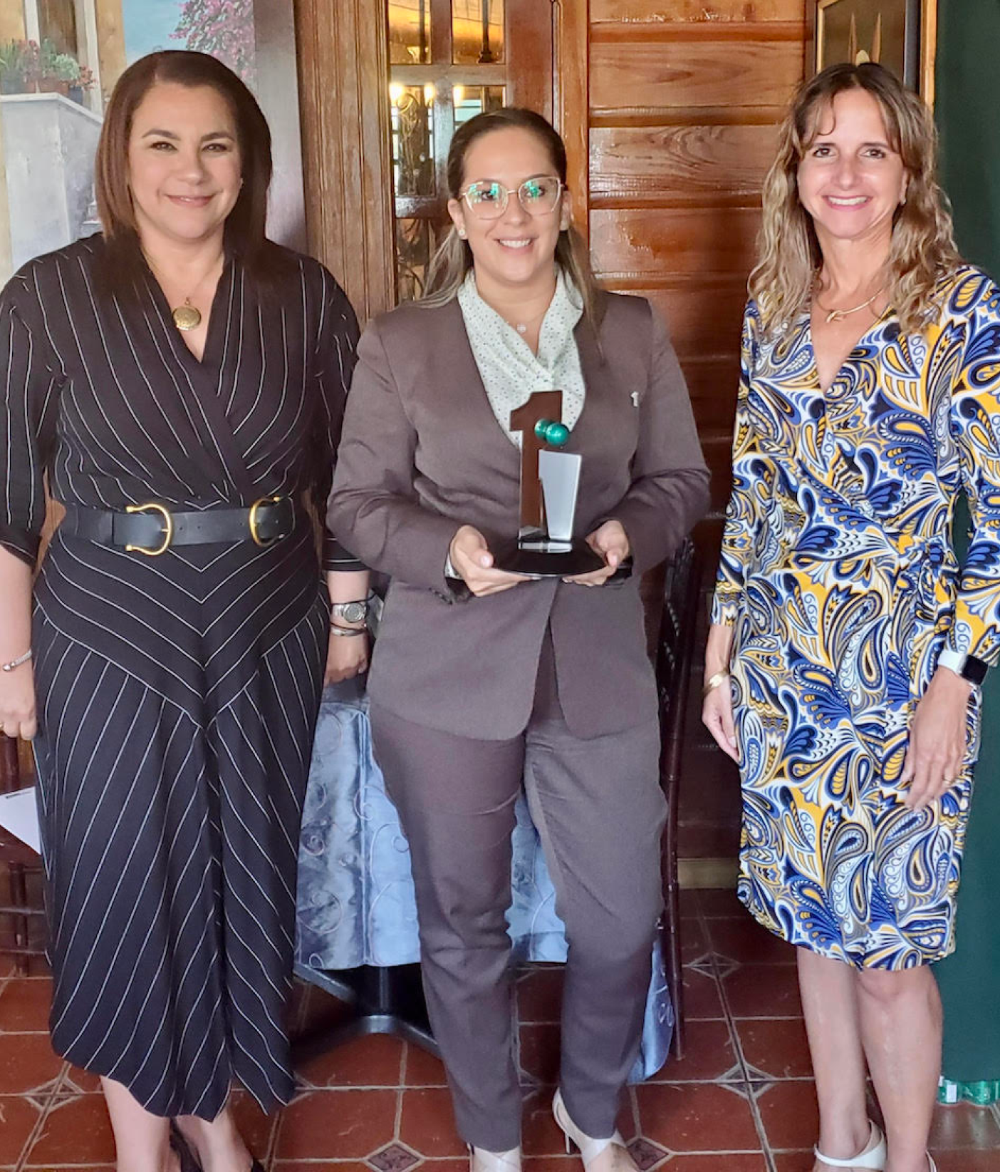 Three women wearing dresses smiling while the one in the middle holds an award