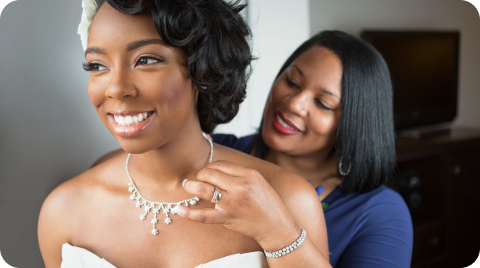 Mother of the bride putting a necklace on the bride while smiling.