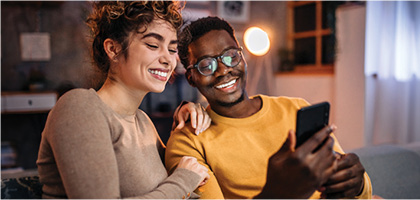 Two people sitting close together on a couch, looking at a smartphone in a warmly lit living room.