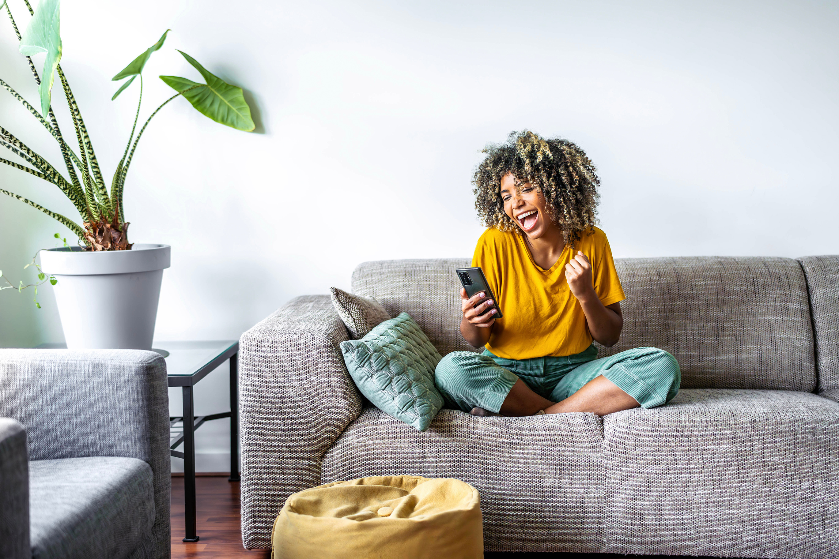 Excited happy young black woman holding smart phone device sitting on sofa at home - Happy satisfied female looking at mobile smartphone screen gesturing yes with clenched fist - Technology concept